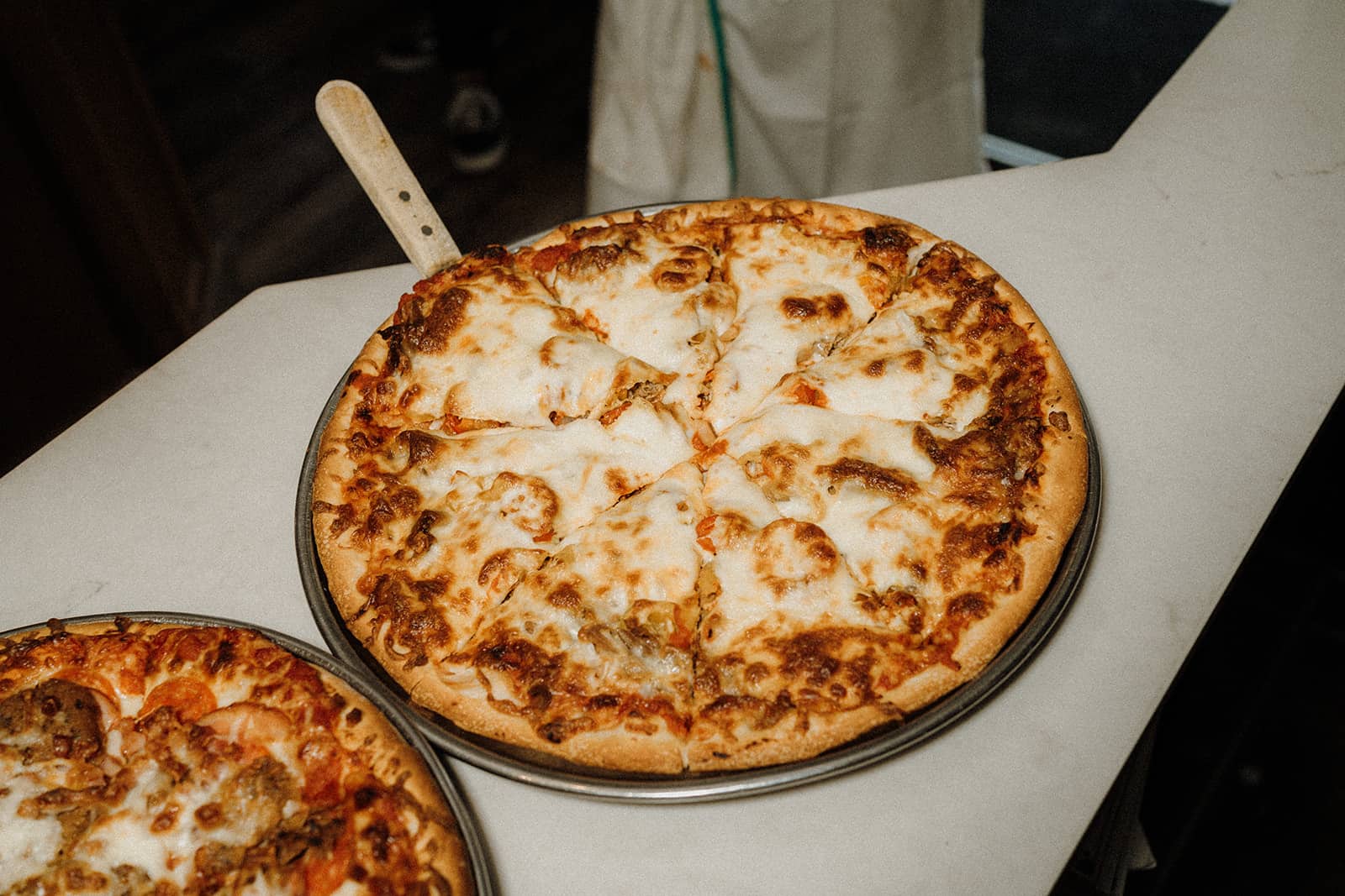 A freshly baked pizza with melted cheese and toppings sits on a metal tray with a handle on a white counter.
