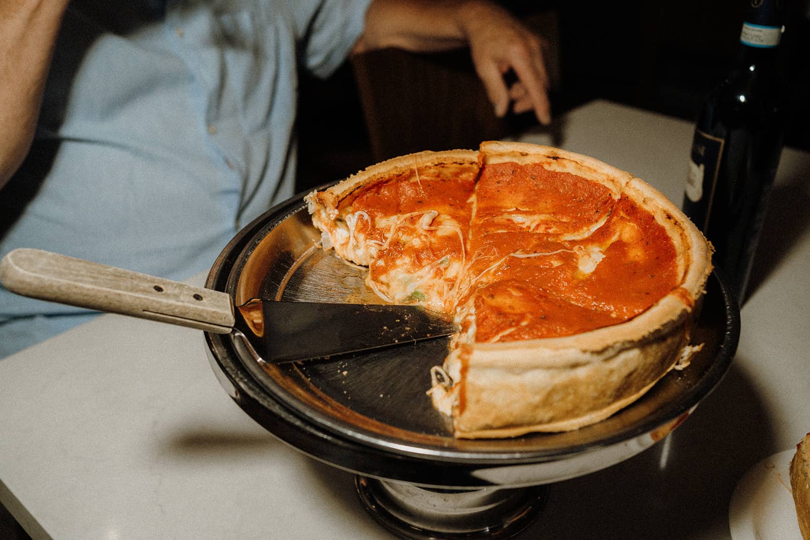 A deep-dish pizza with two slices missing sits on a metal stand next to a pizza server and a bottle on the table.