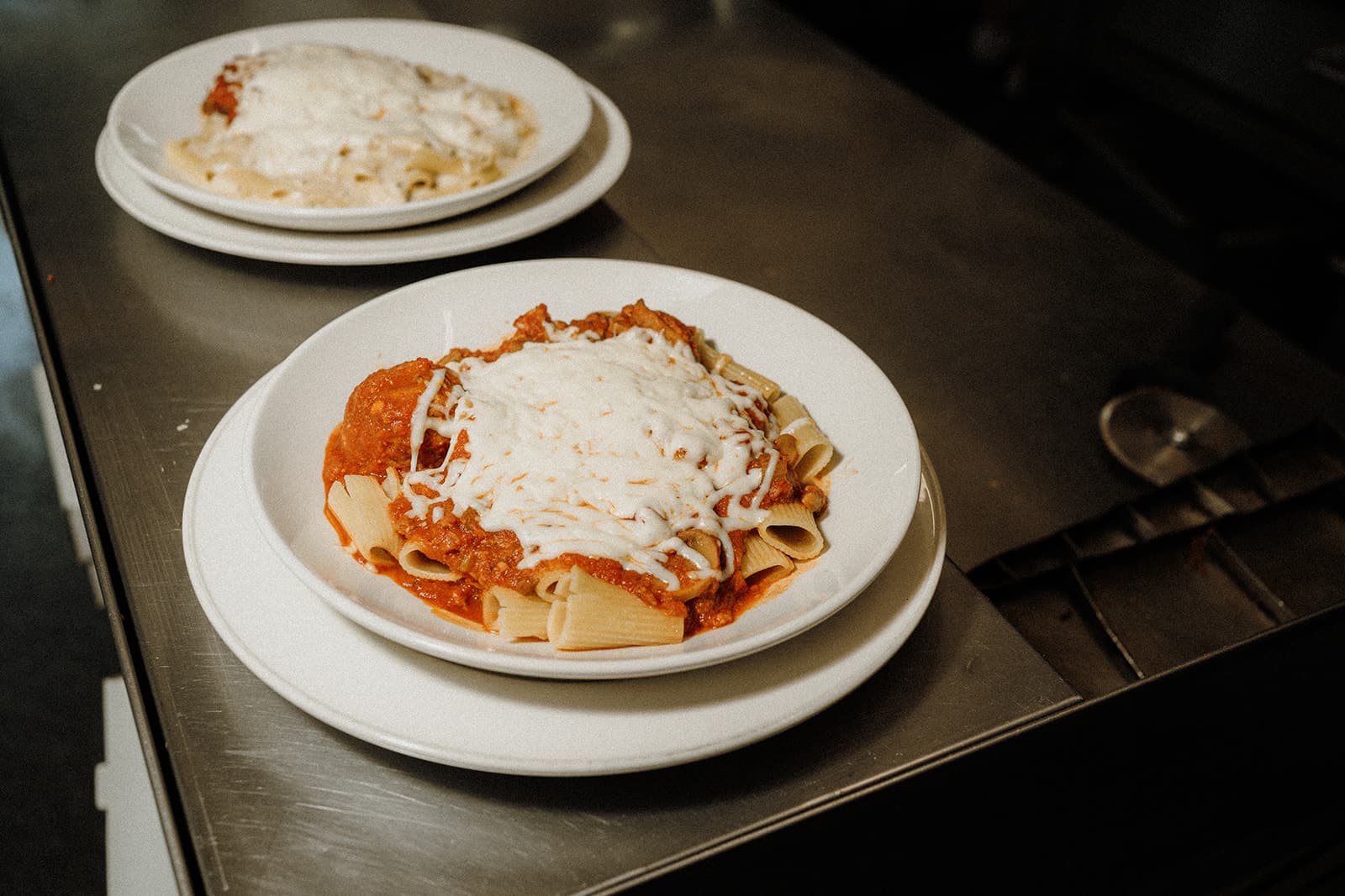 Two plates of pasta topped with tomato sauce and shredded cheese on a stainless steel surface.