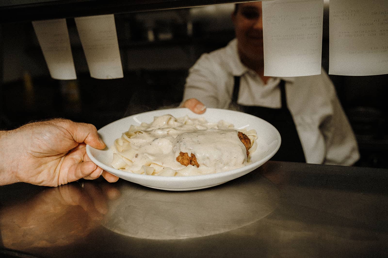 A server hands a plate of pasta with creamy white sauce and breaded meat across a restaurant counter.