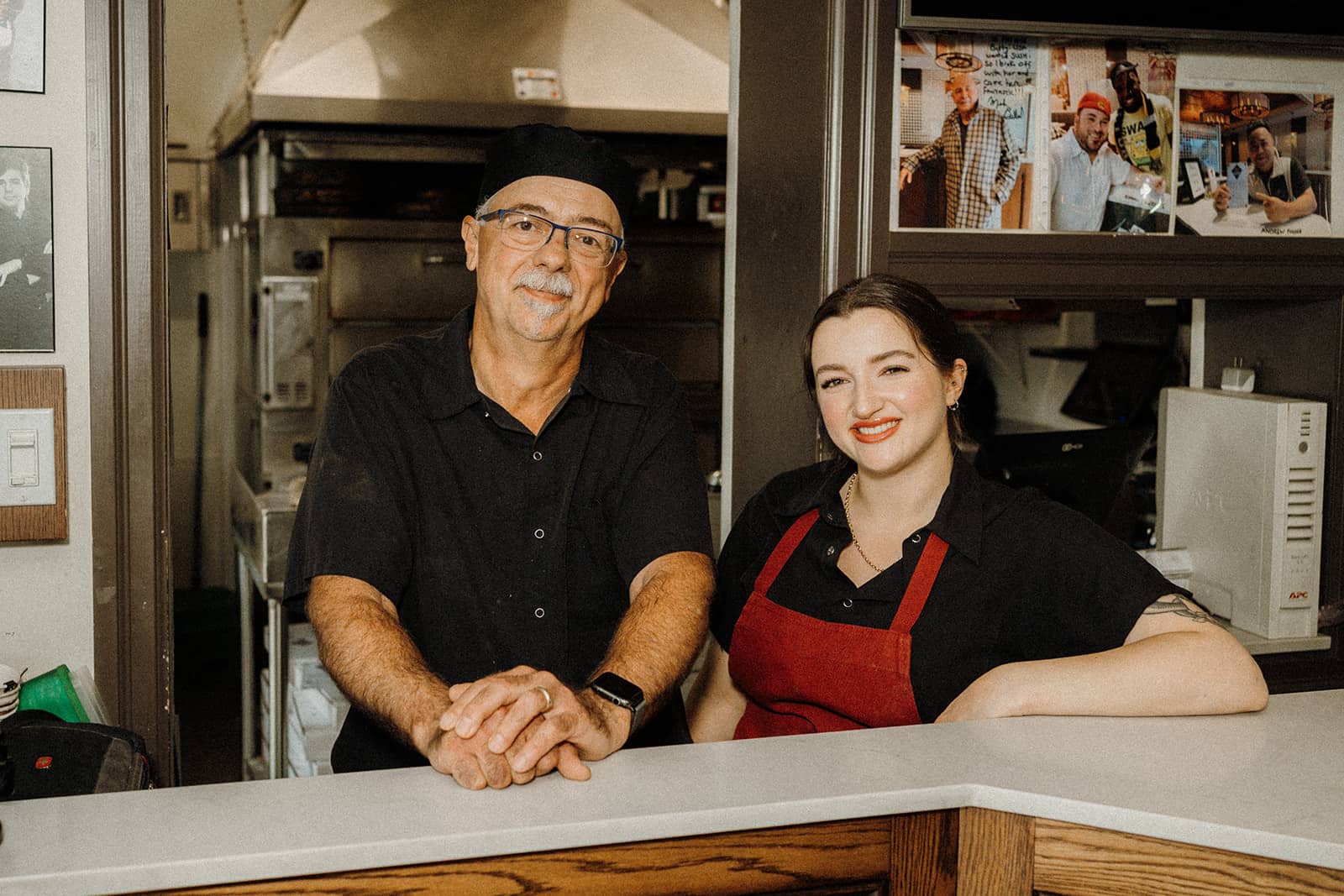 Two people in black shirts stand behind a counter in a kitchen, smiling at the camera.