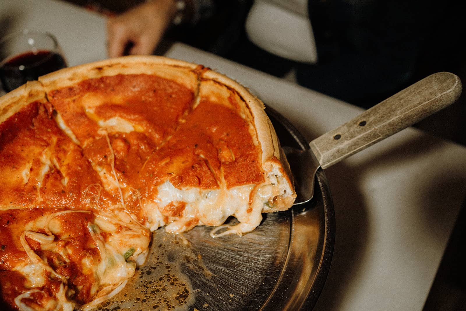 A deep-dish pizza with melted cheese and tomato sauce sits on a metal tray with a server under one slice.