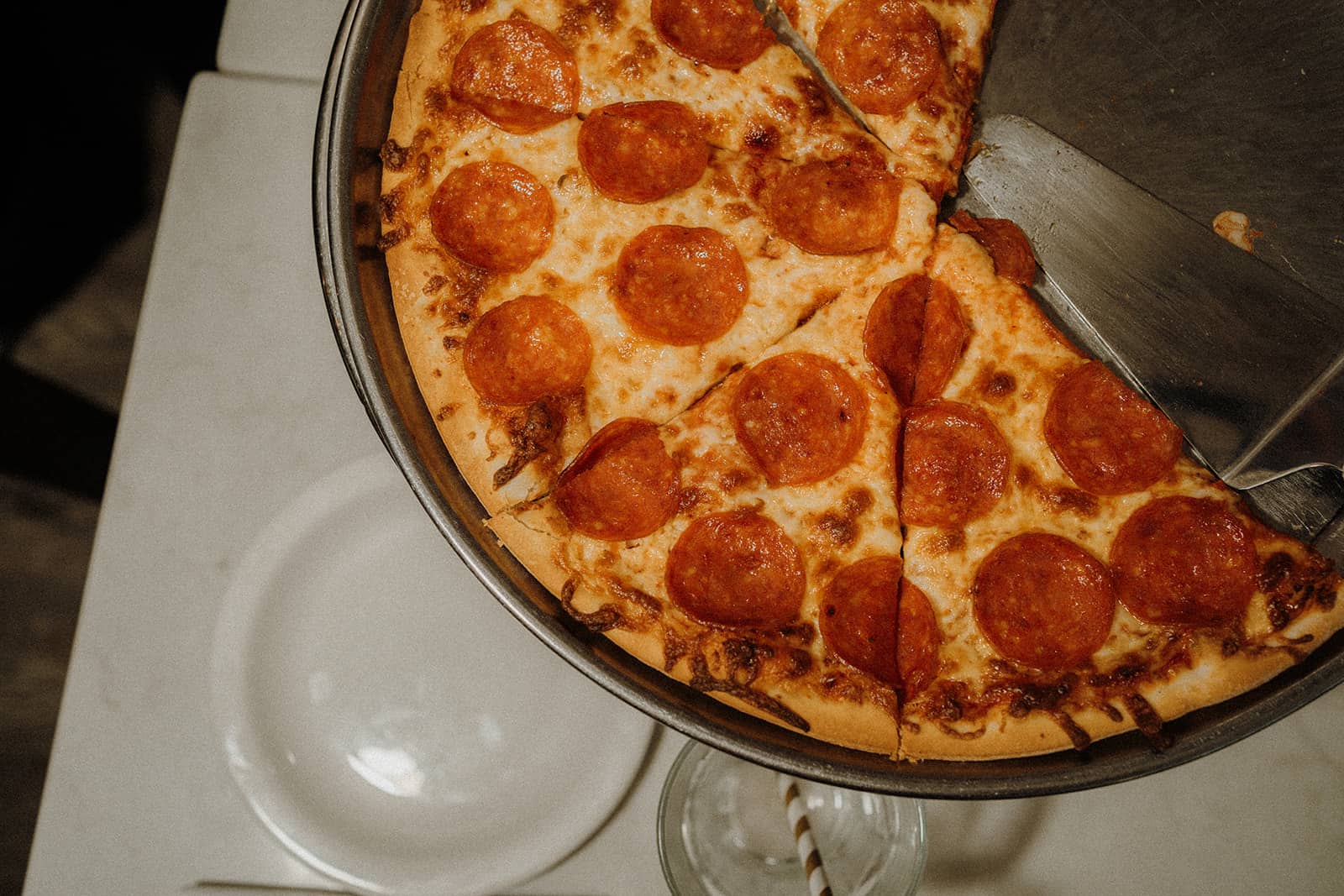 A pepperoni pizza with several slices missing sits on a metal tray above a white plate and a glass with a straw.