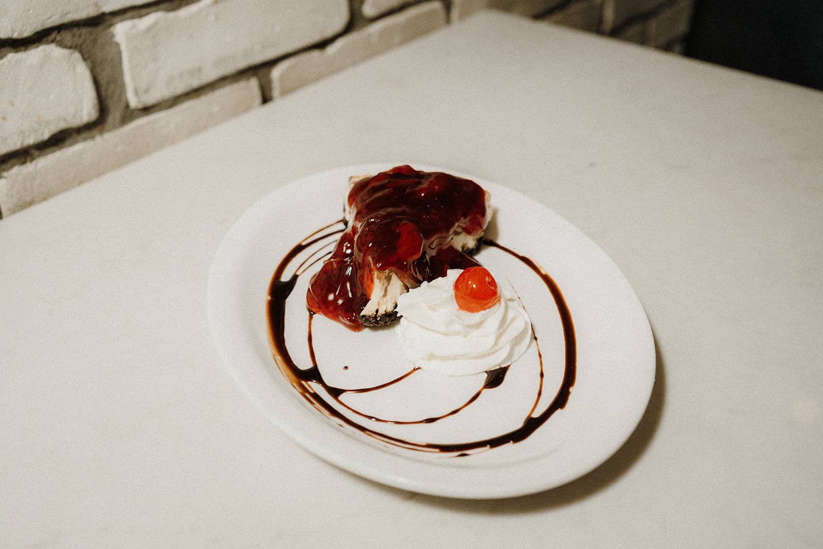 Slice of cheesecake with cherry topping, chocolate drizzle, whipped cream, and a cherry on a white plate.