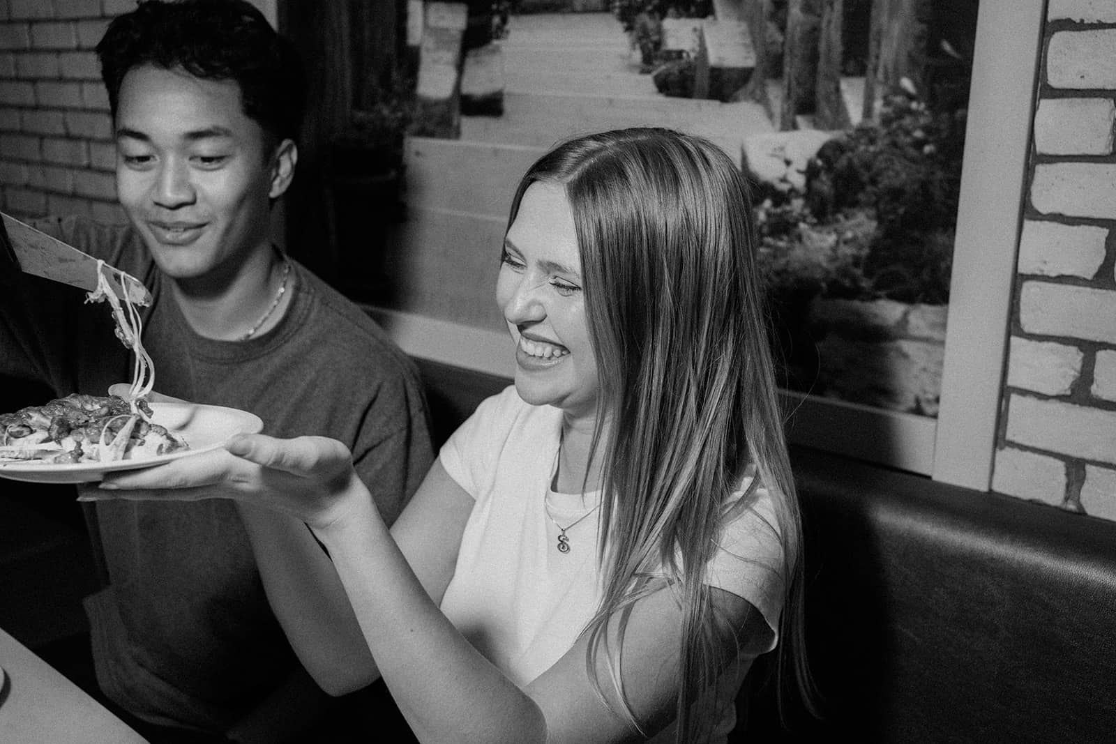 Two people sit together as one of them lifts a slice of pizza from a plate, smiling.