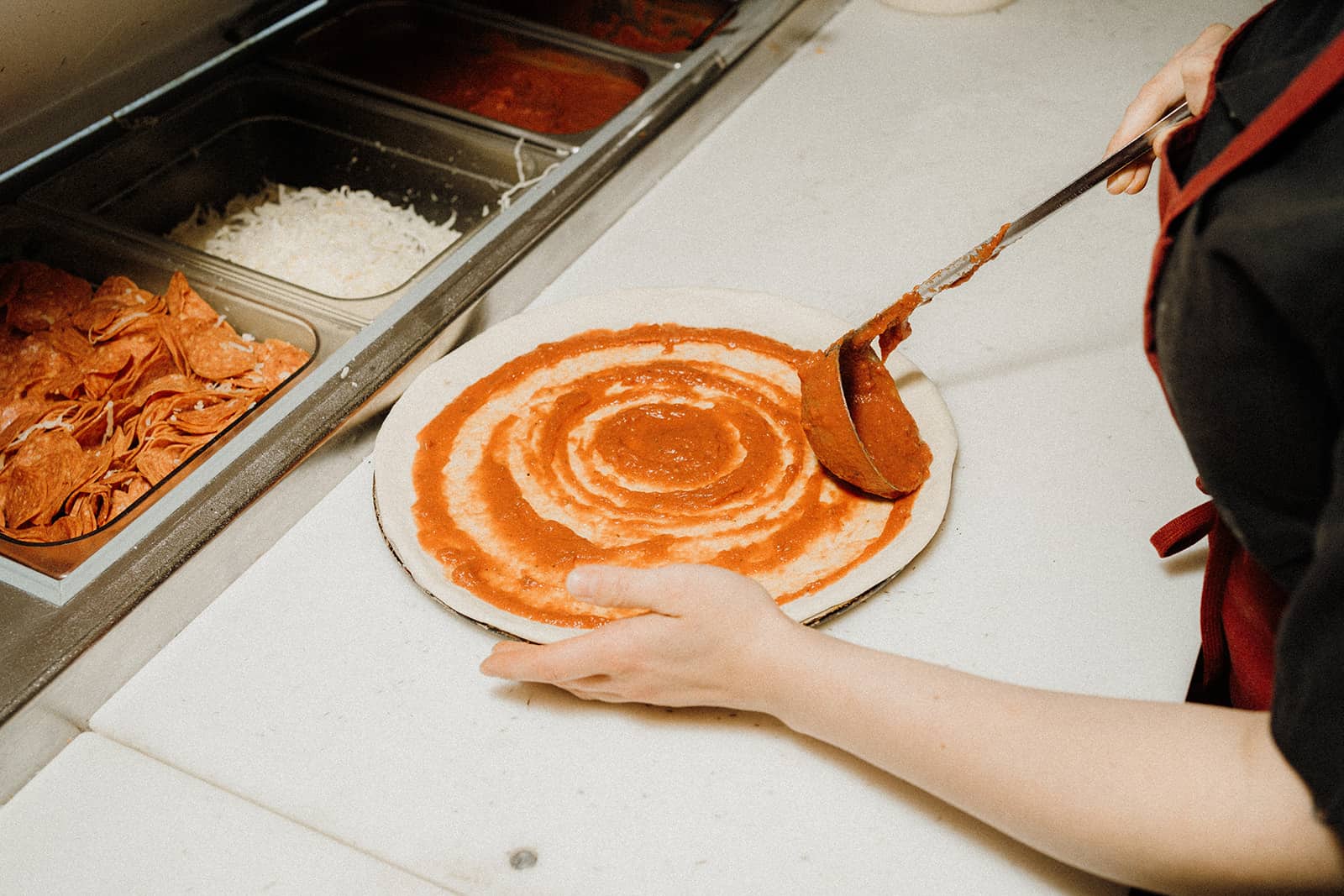 A person spreads tomato sauce on pizza dough at a prep station with toppings and cheese nearby.