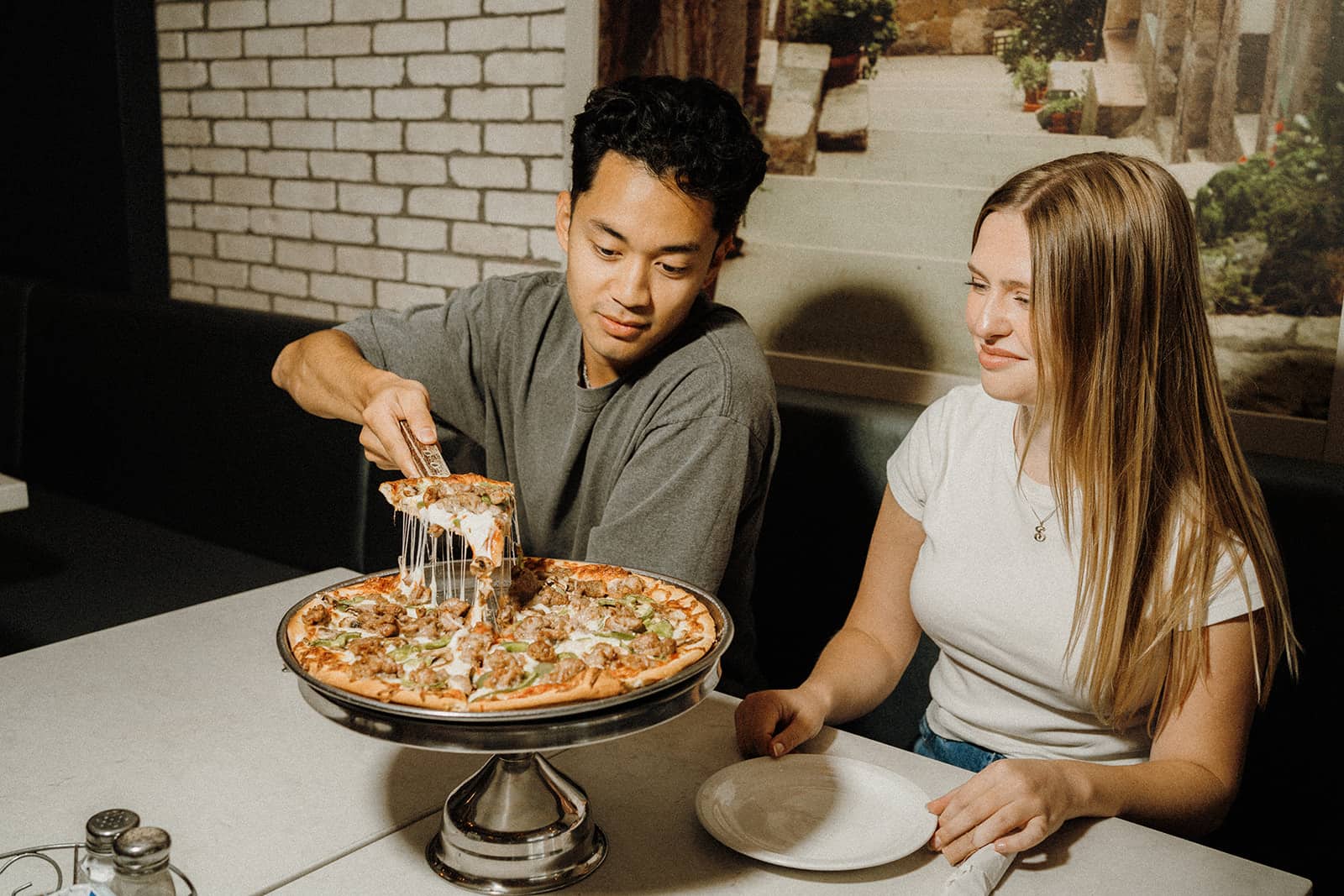 A man serves a slice of pizza from a stand while a woman sits beside him holding an empty plate.