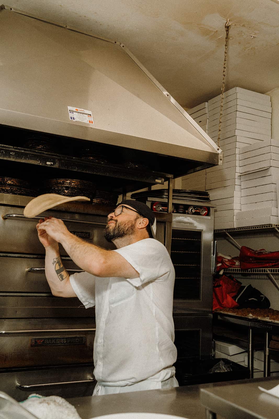 A man in a white uniform tosses pizza dough in a commercial kitchen with stacked pizza boxes in the background.