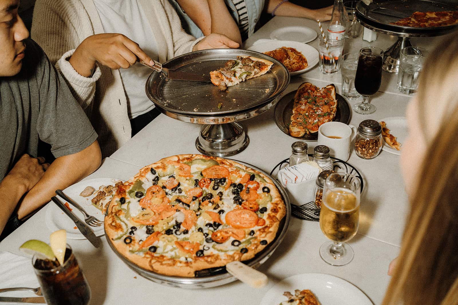 People seated at a table share pizza, drinks, and other dishes at a casual restaurant meal.