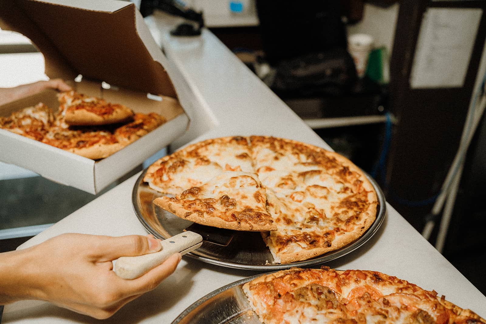 A hand cuts a pizza on a tray while another pizza is being placed into a takeout box on a counter.