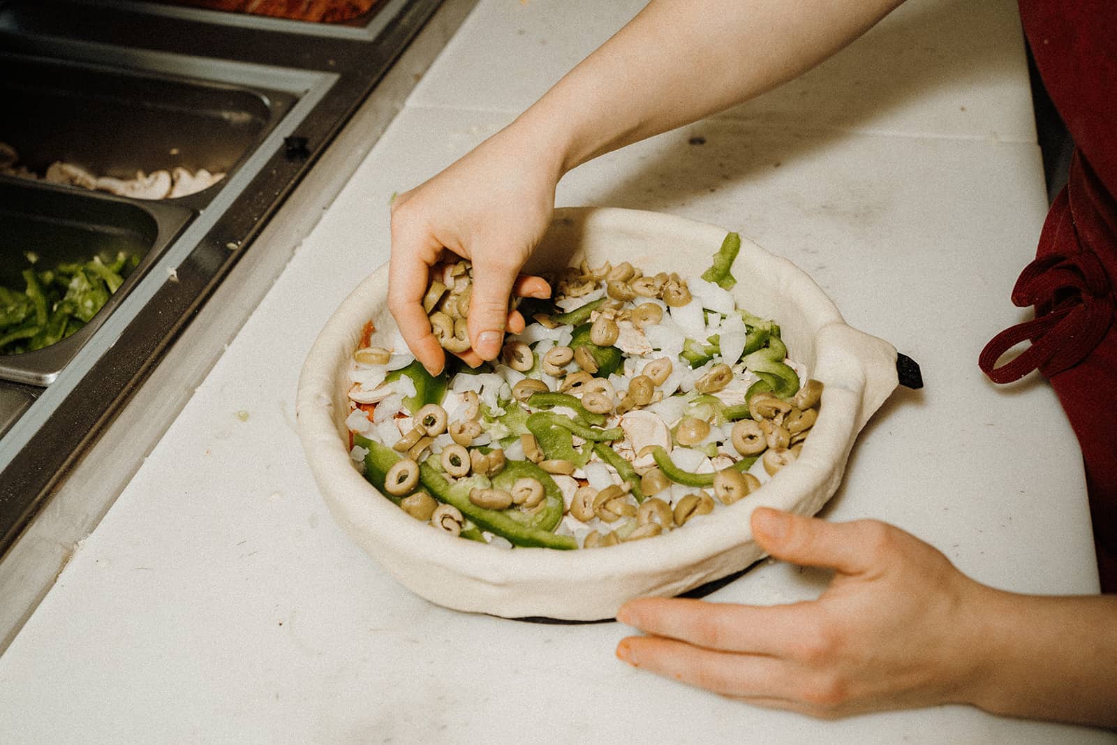 A person adds sliced green olives to a deep-dish pizza crust filled with vegetables on a prep counter.