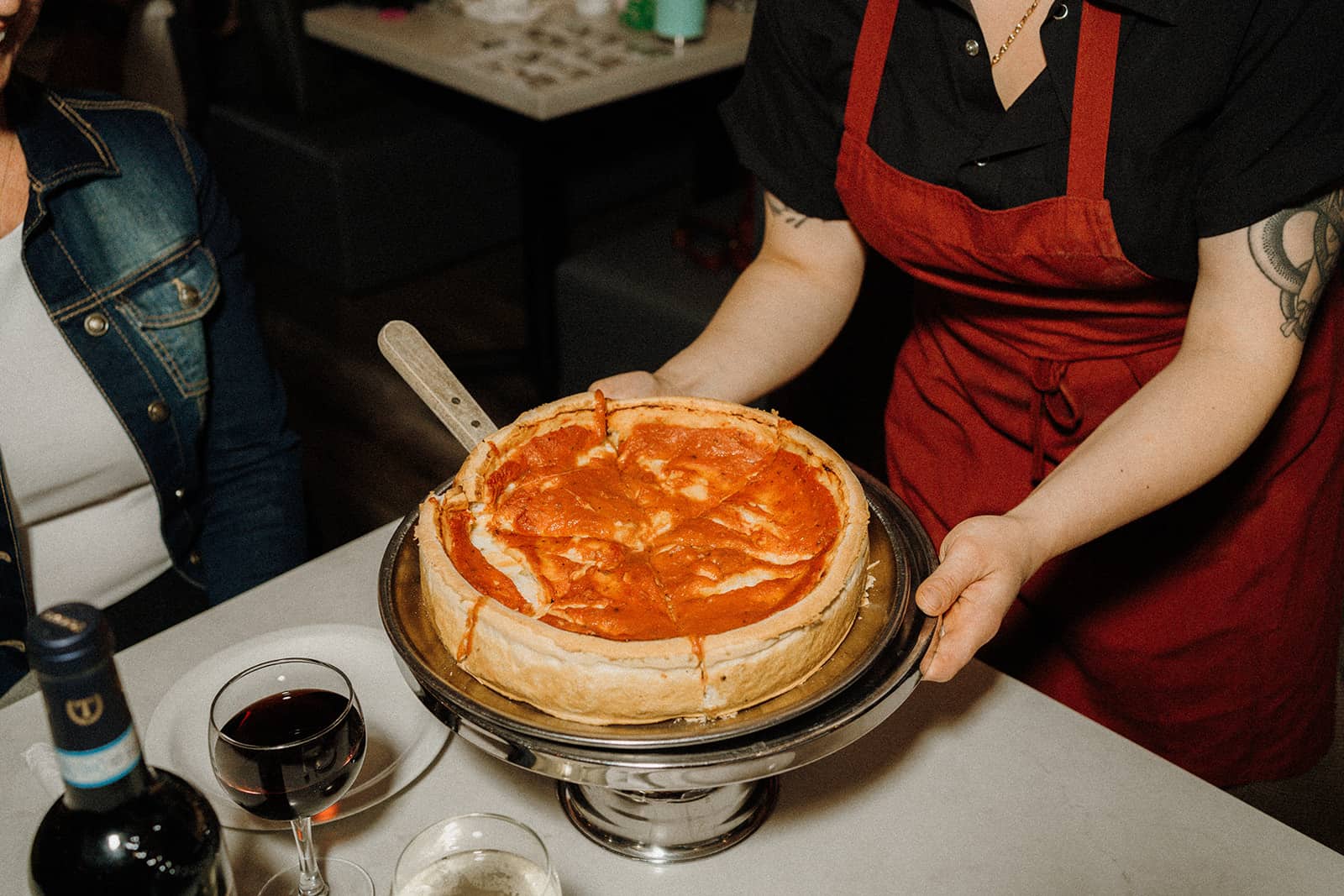 A person in a red apron serves a deep-dish pizza on a metal stand at a table with drinks.