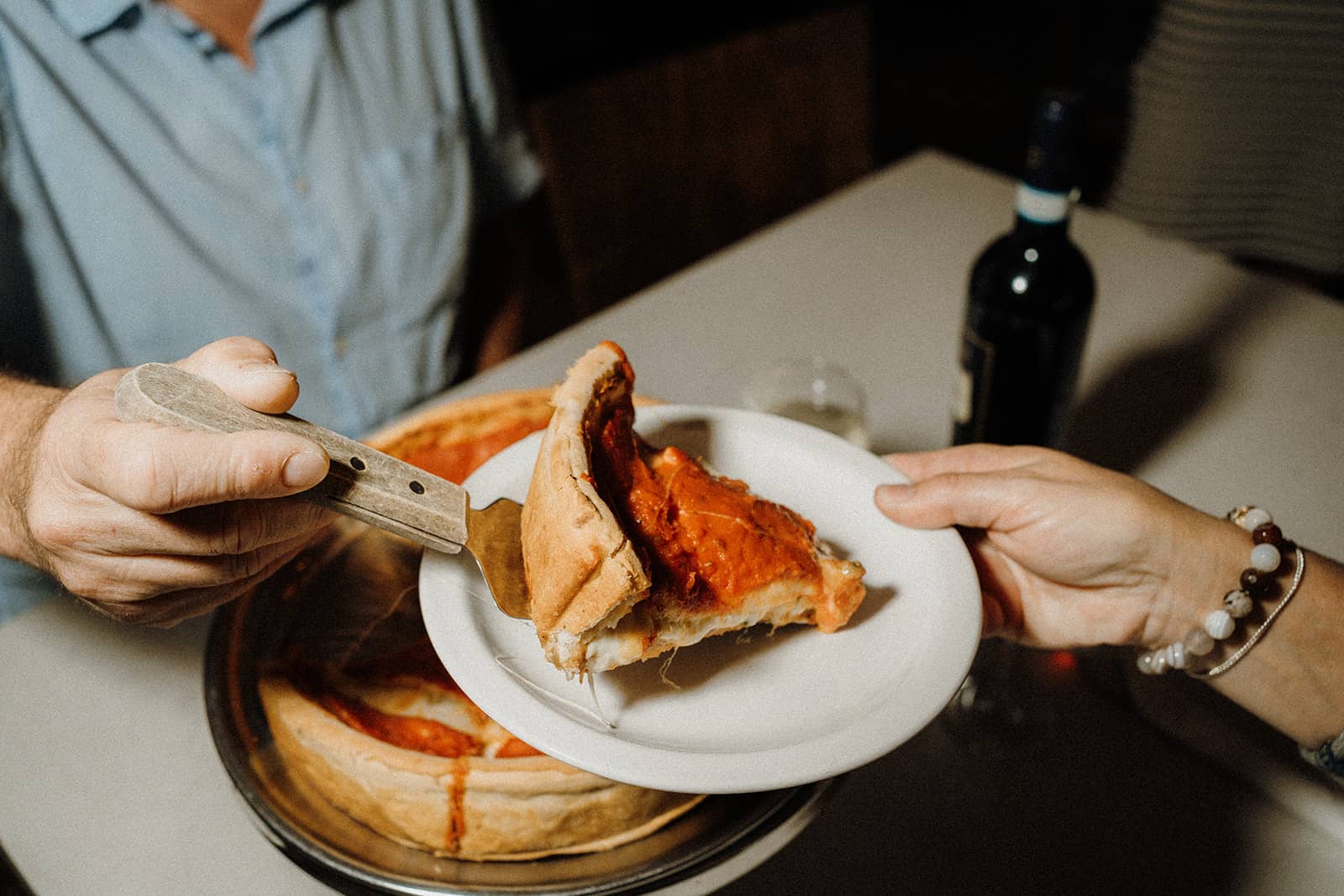 A hand serves a slice of deep-dish pizza onto a plate held by another person at a table with wine.