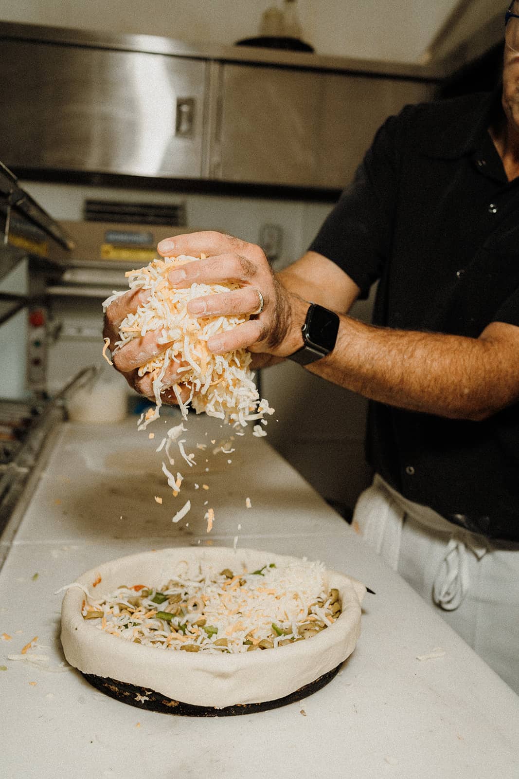 A person sprinkles shredded cheese onto an uncooked pizza crust in a kitchen setting.