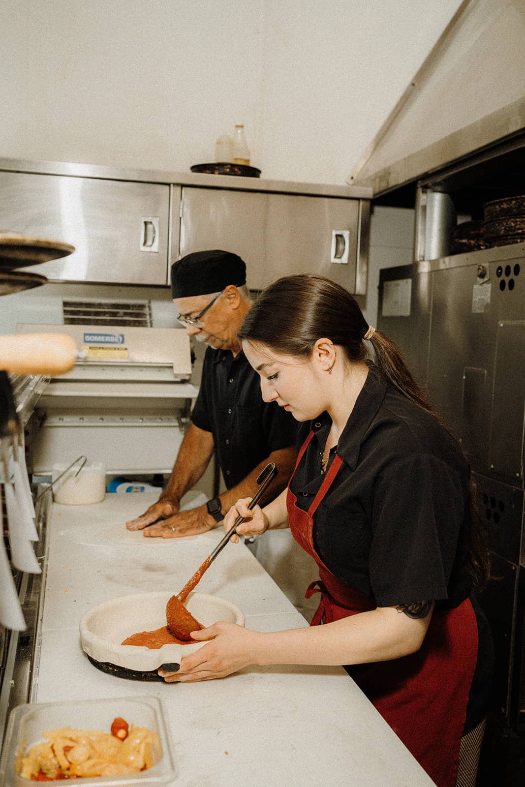 Two people preparing pizza in a kitchen; one spreads sauce on dough while the other works nearby.