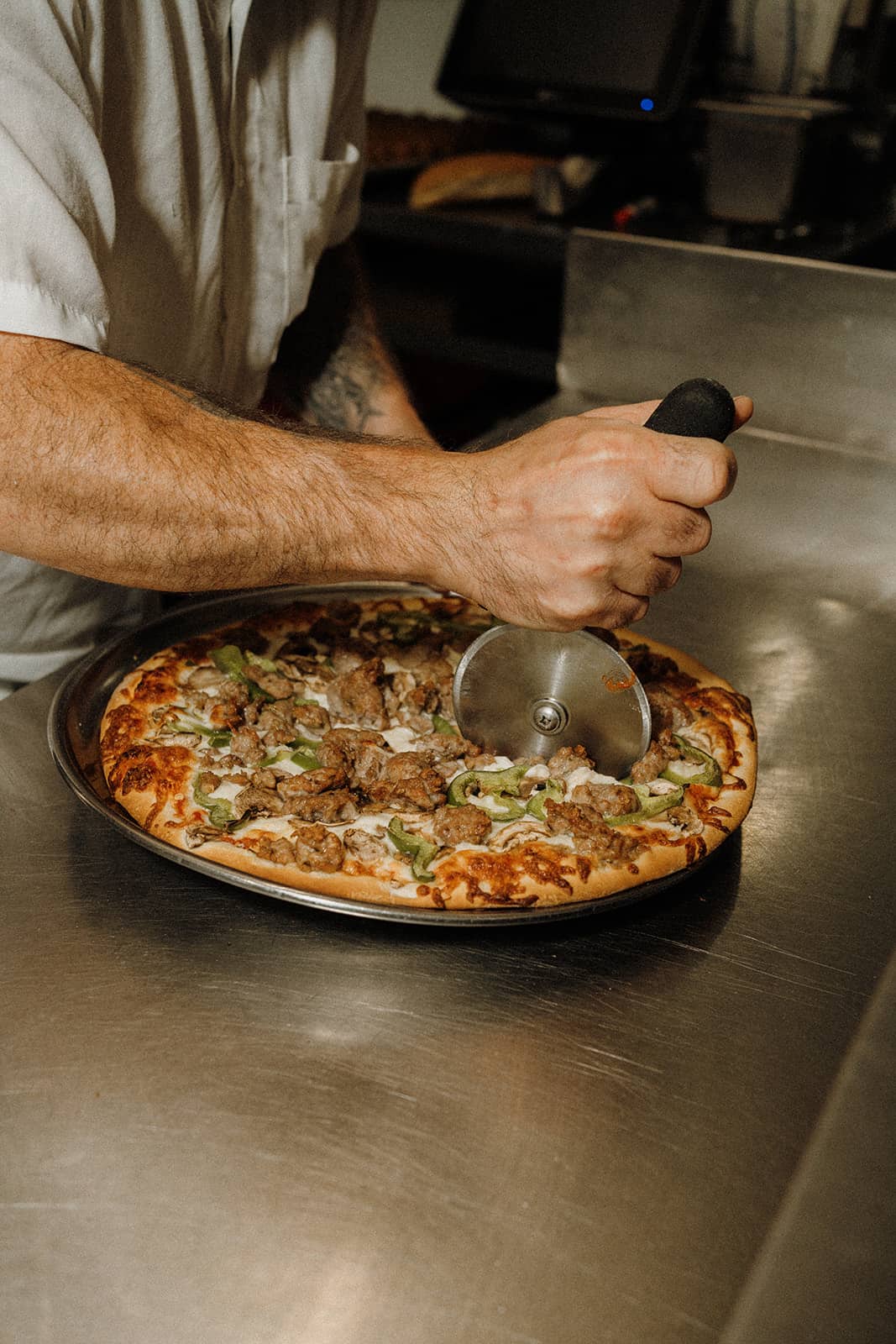 A person slices a cooked pizza with a pizza cutter on a metal counter.