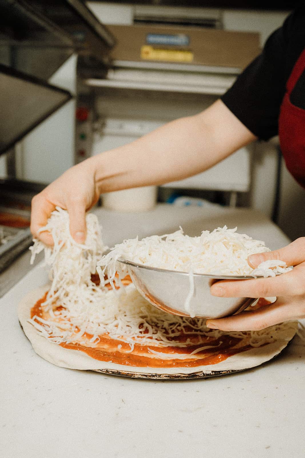 A person sprinkles shredded cheese from a bowl onto pizza dough covered with tomato sauce.