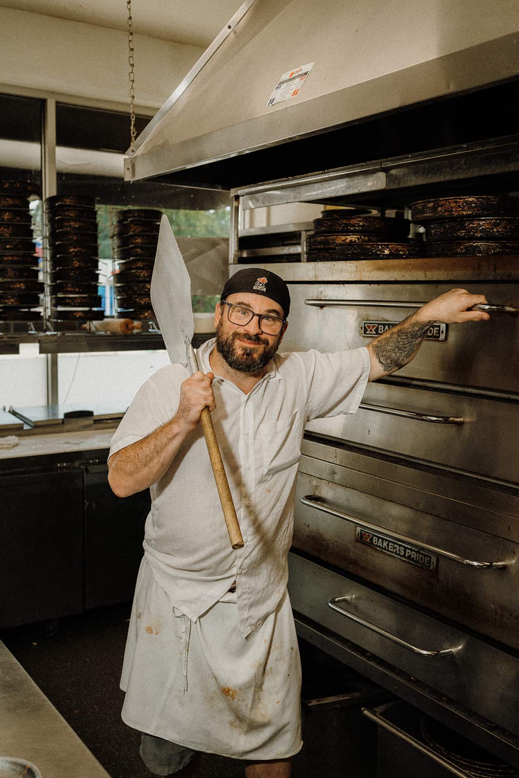 A man in a white apron and cap stands by an industrial oven, holding a pizza peel in a commercial kitchen.
