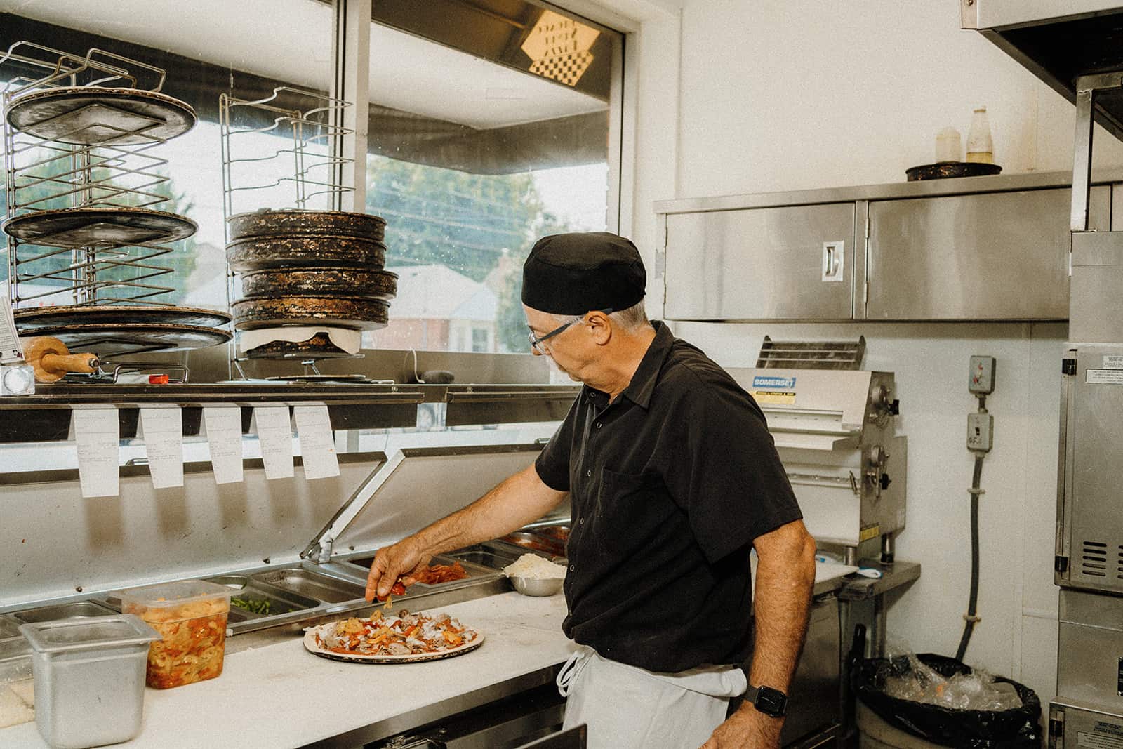 A man in a black shirt and hat prepares a pizza at a counter in a commercial kitchen.