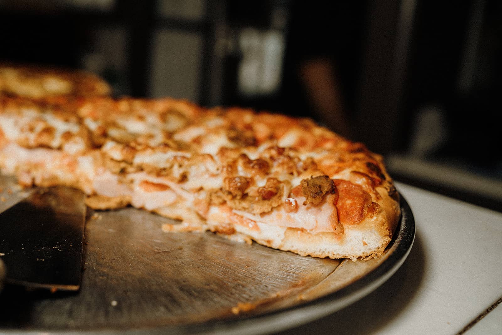 A partially sliced pizza with meat toppings sits on a metal tray, with a spatula next to it.