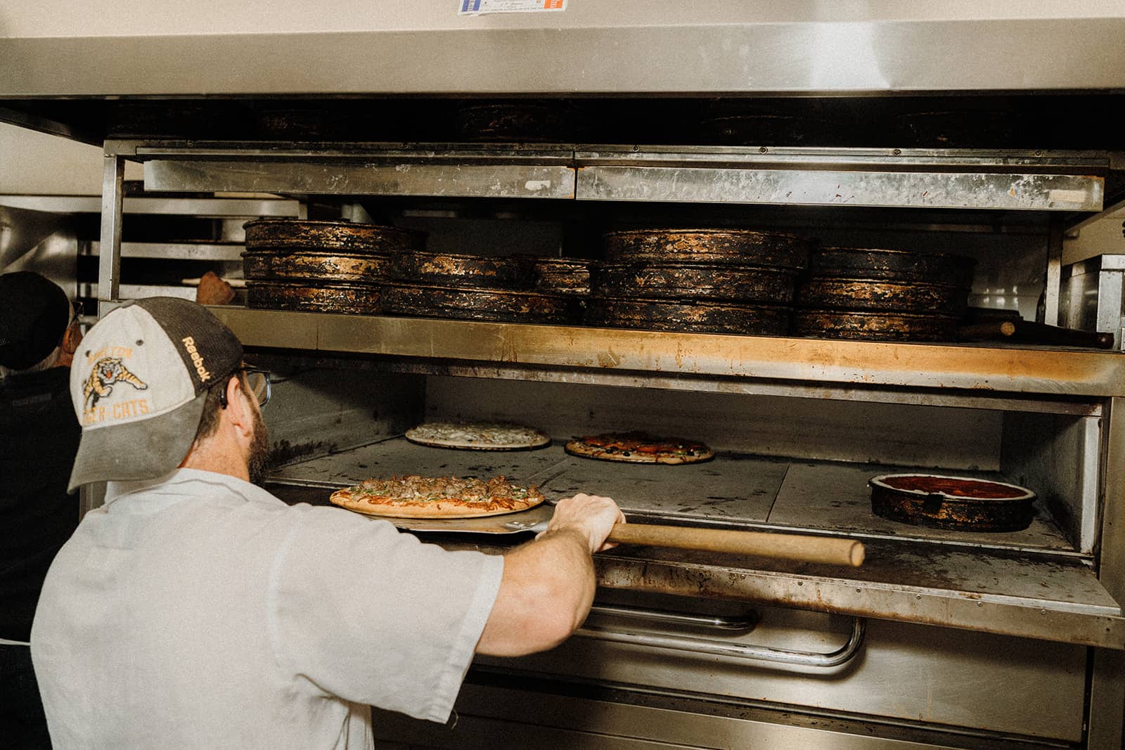Person placing a pizza into a commercial oven with several pizza pans stacked on top of the oven.