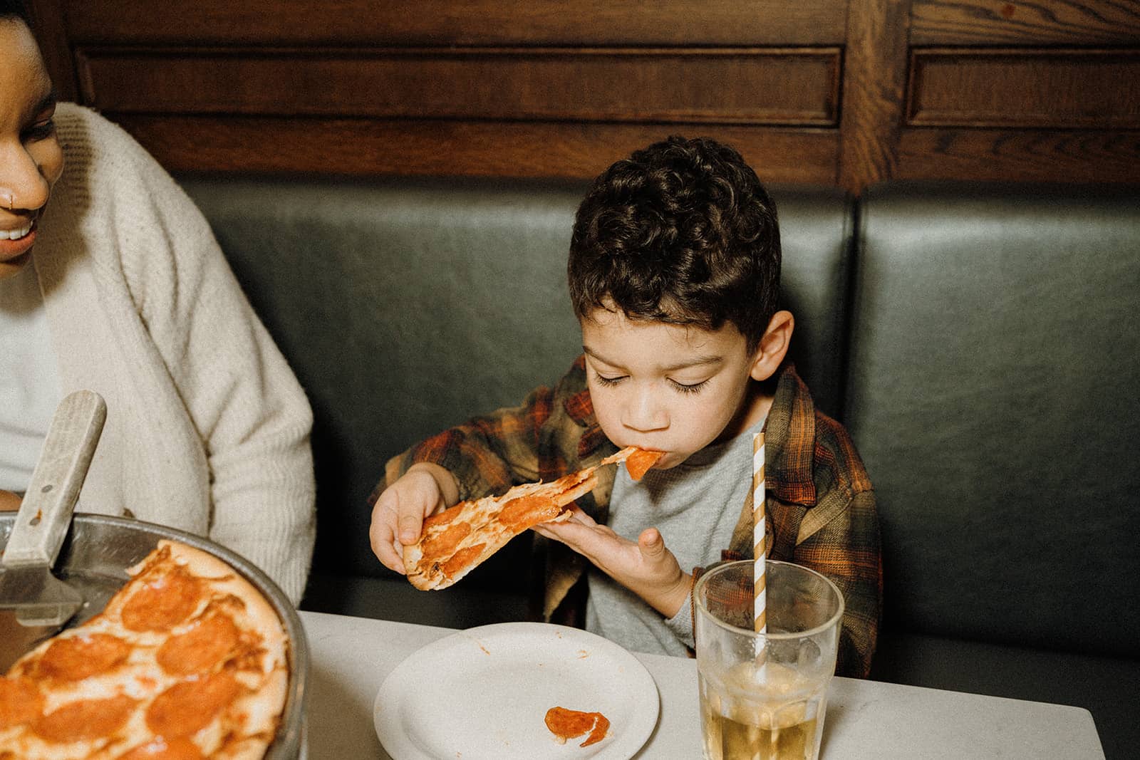 A young boy eats a slice of pepperoni pizza at a restaurant table with a drink and an adult nearby.