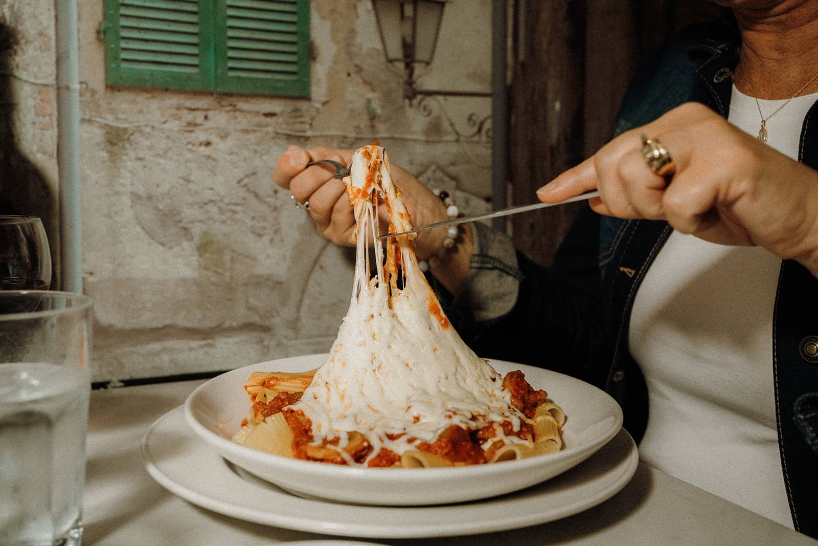 A person lifts a forkful of cheesy baked pasta from a plate at a restaurant table.