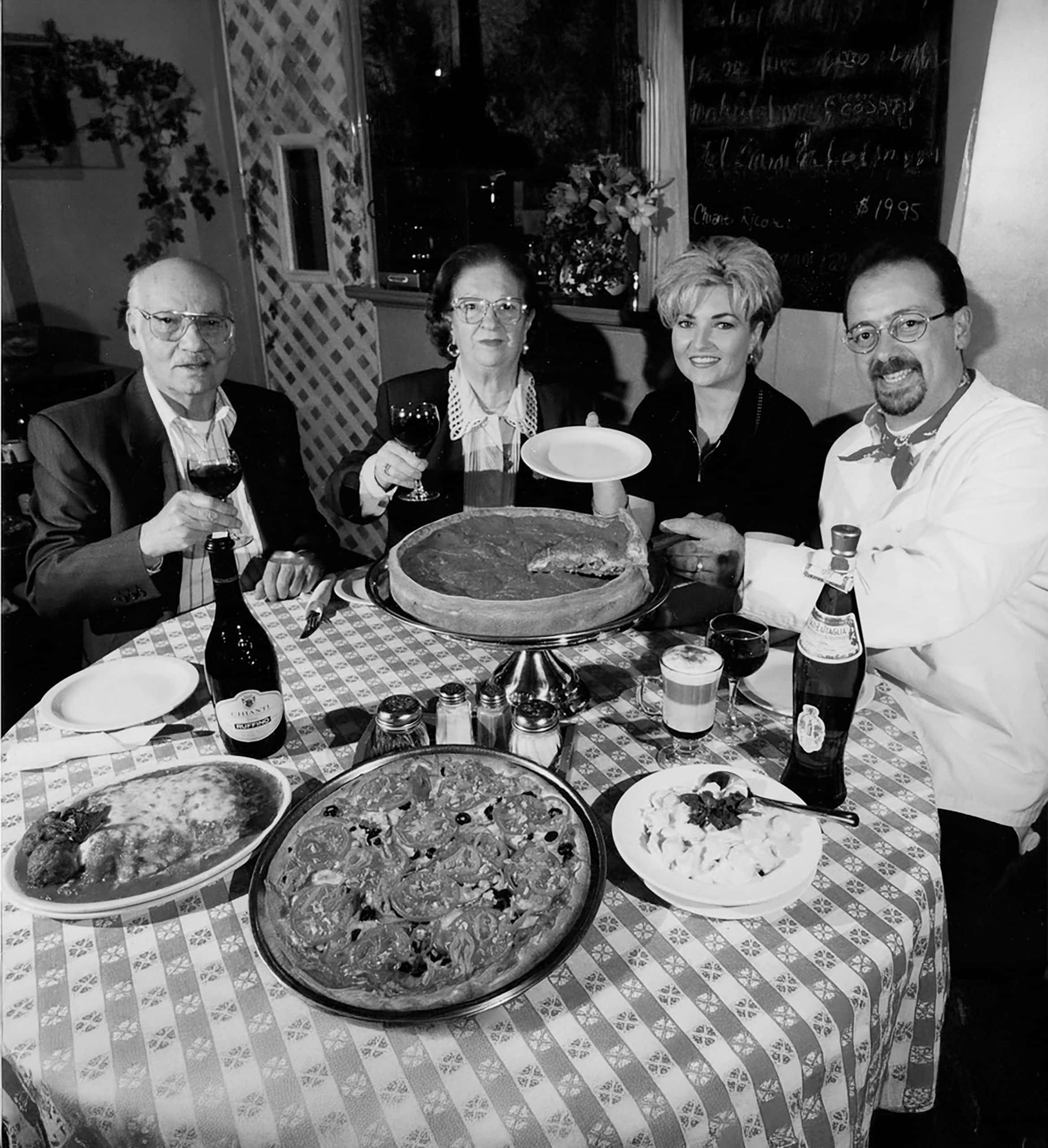 Four adults sit at a round table with pizza and drinks, raising glasses and smiling for the camera in a restaurant.