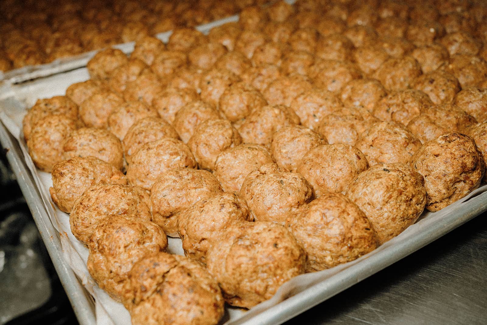 Rows of baked meatballs arranged closely on a parchment-lined baking sheet in a kitchen setting.