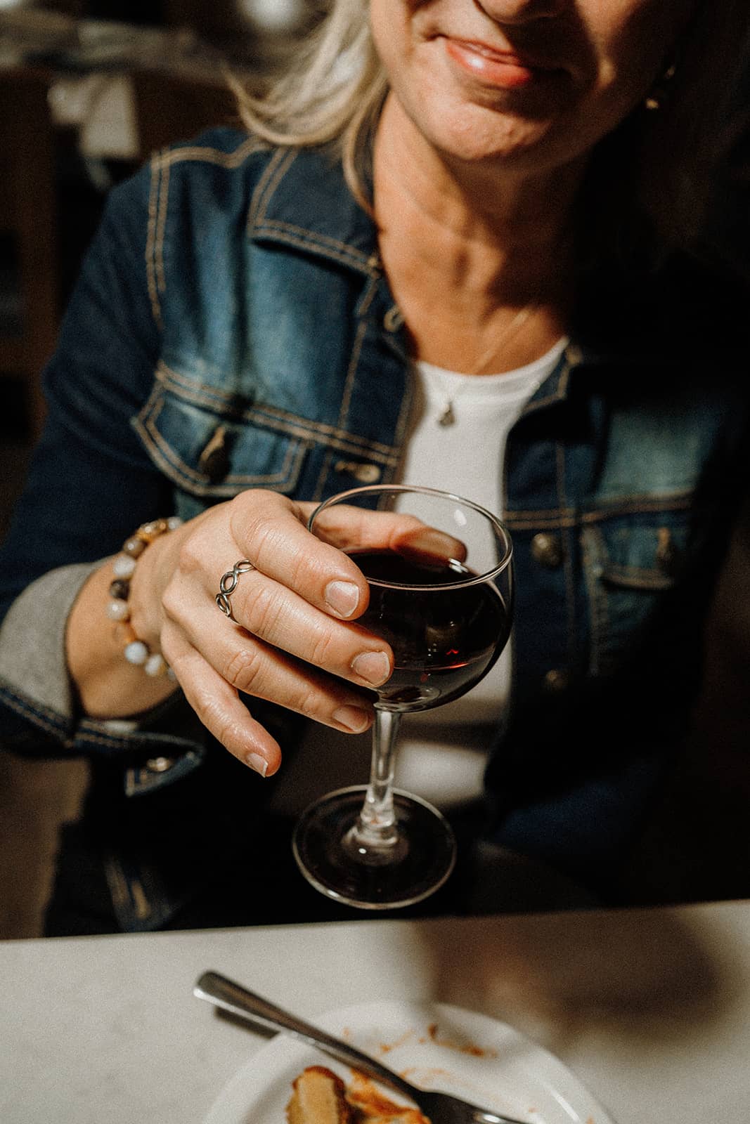A woman in a denim jacket holds a glass of red wine at a table with a partially eaten dessert.