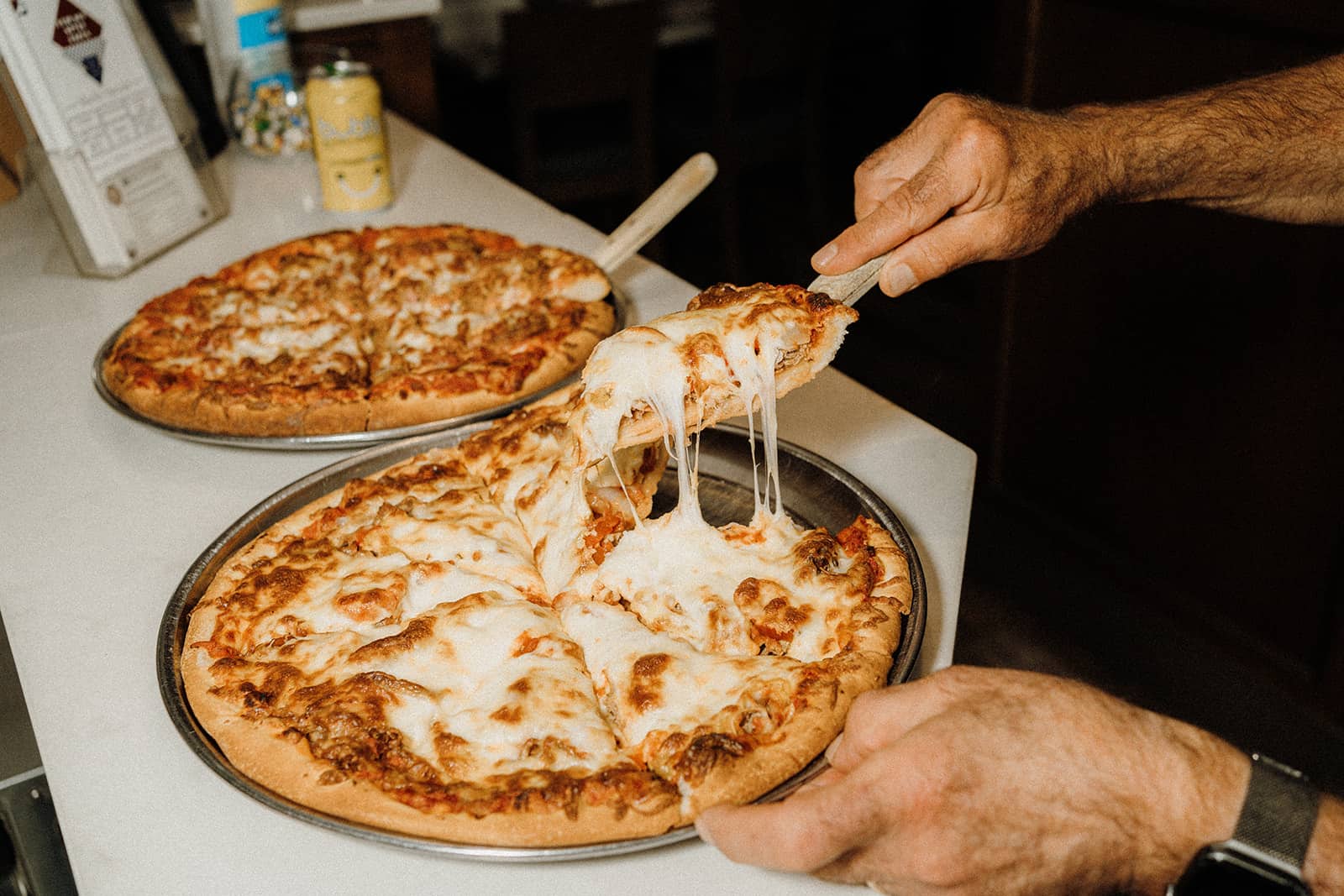 A person lifts a cheesy slice from a baked pizza on a metal tray; another whole pizza sits in the background.