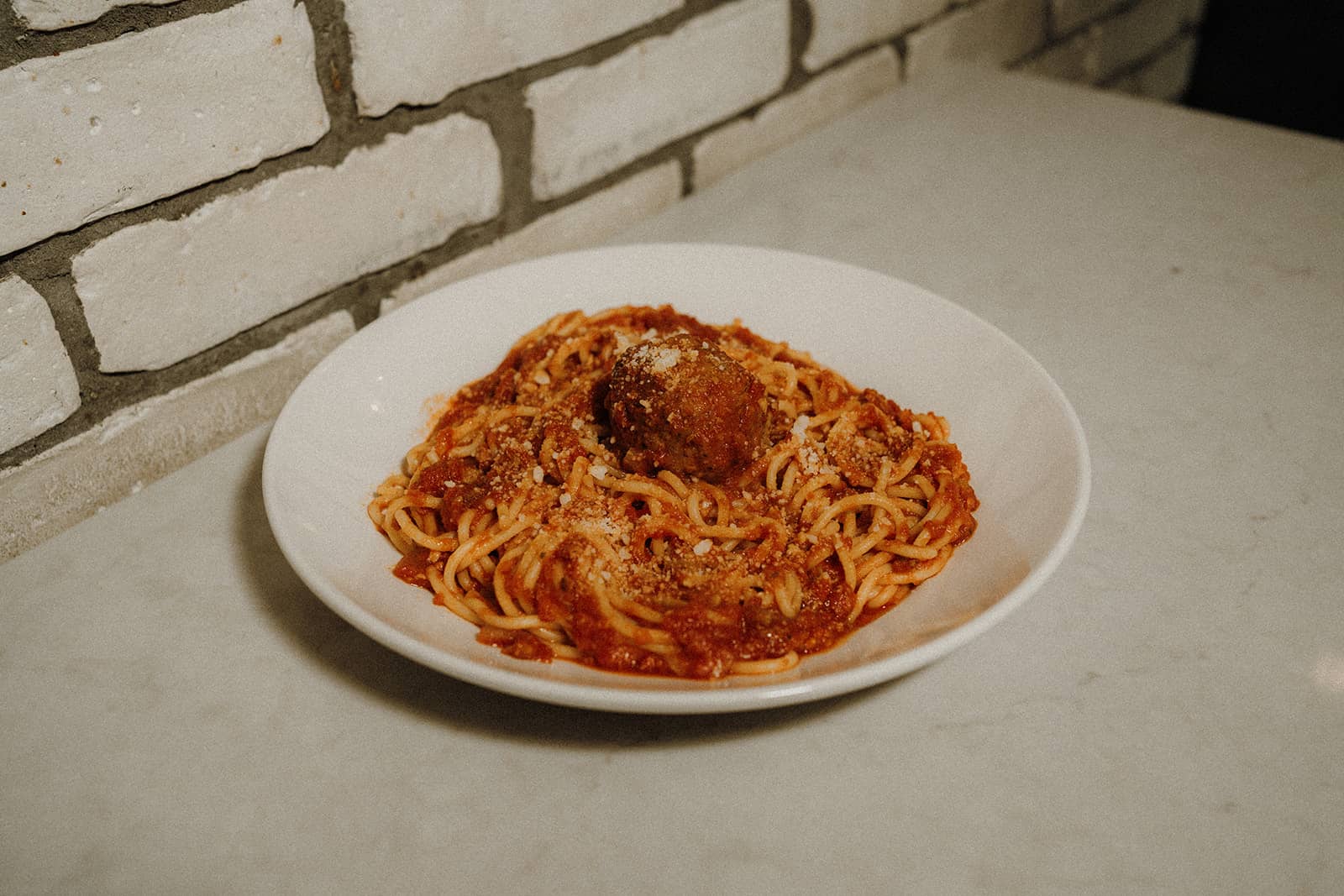 A plate of spaghetti with marinara sauce and a meatball, sprinkled with grated cheese, on a marble countertop.