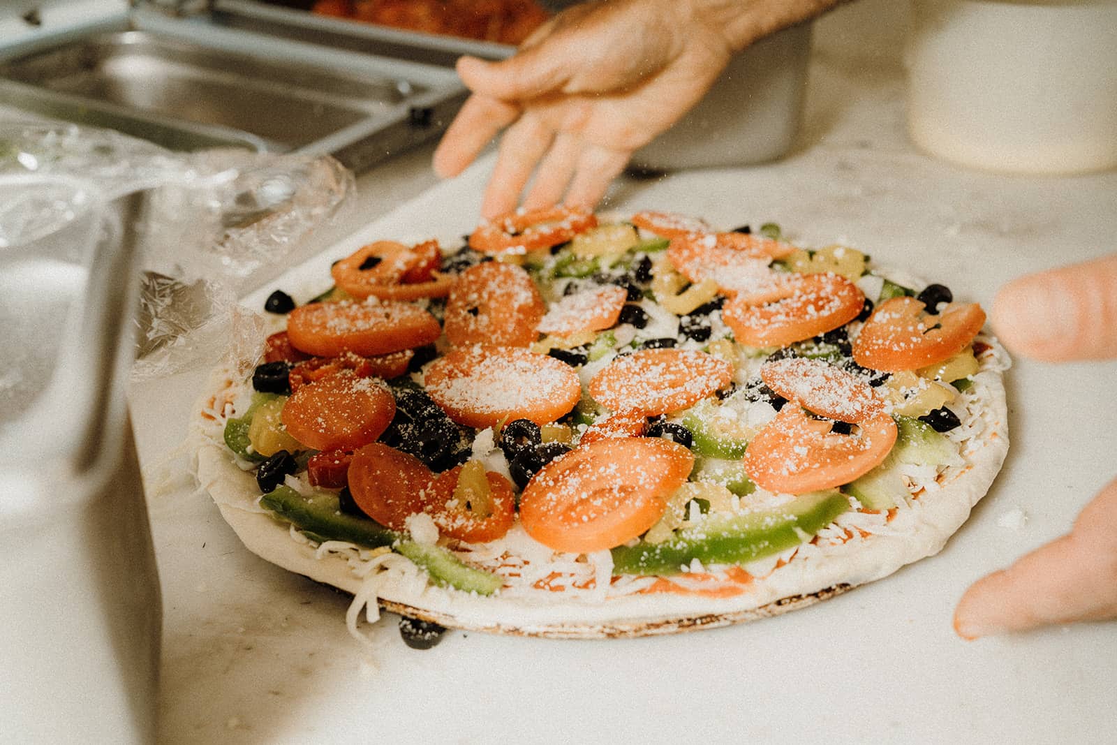 A person prepares a pizza topped with tomato slices, green peppers, olives, and cheese on a kitchen counter.