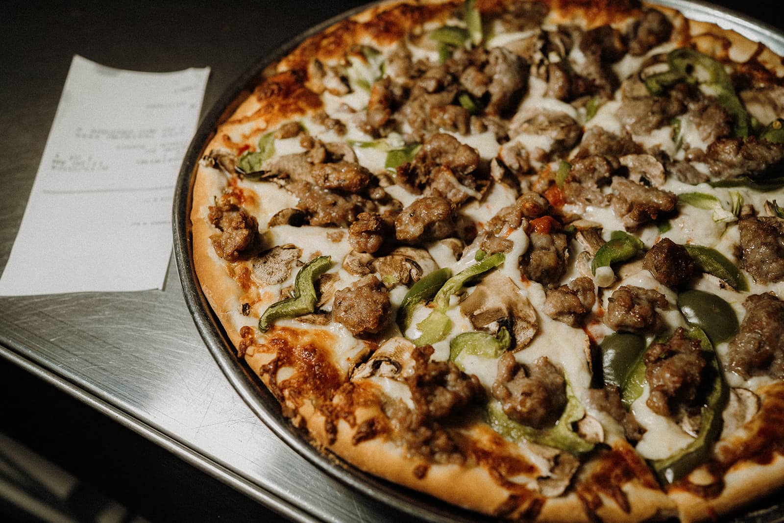 A baked pizza with sausage and green peppers sits on a tray next to a printed receipt on a metal surface.
