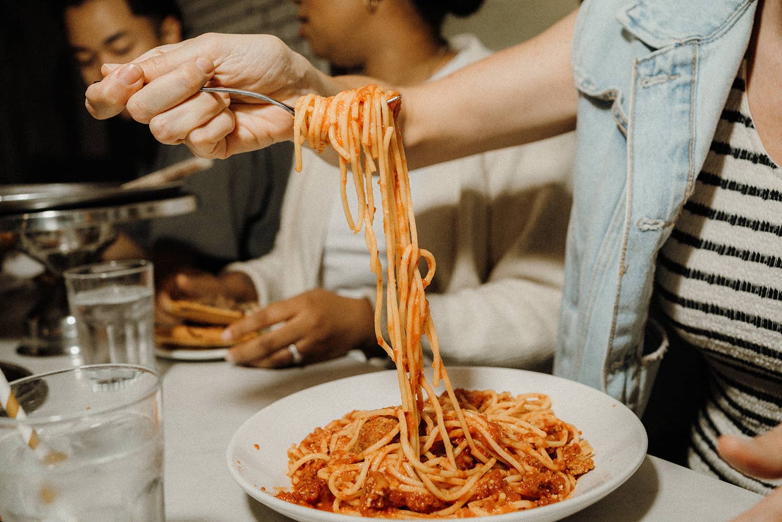 Person twirling spaghetti with tomato sauce on a fork above a plate, seated at a table with others and glasses of water.